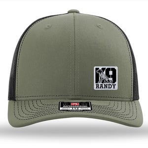 May include: An olive green baseball cap with black mesh back panels and a structured front. A white square patch on the front features a black silhouette of a dog, the text K9, and the name RANDY. A black sticker is on the brim with white text.