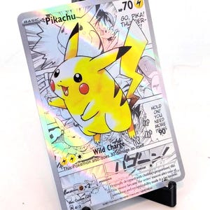 Puede incluir: Una tarjeta coleccionable holográfica de Pikachu, con un personaje Pikachu amarillo con detalles en negro. La tarjeta muestra el texto "Wild Charge" y "-70". La tarjeta se muestra en un soporte negro.