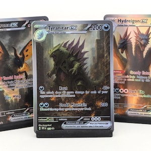 May include: Three Pokemon EX trading cards are displayed. The cards feature Volcarona, Tyranitar, and Hydreigon. The Volcarona card shows a large insect with orange and black wings. The Tyranitar card depicts a green, spiky monster. The Hydreigon card shows a three-headed dragon.