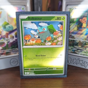Puede incluir: Una tarjeta Pokémon de Bulbasaur enmarcada con un diseño 3D en capas. La tarjeta presenta un fondo verde y blanco con el personaje de Bulbasaur en el centro. El marco es de color azul oscuro y la tarjeta se exhibe en una estantería de madera.
