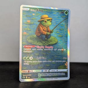 May include: A holographic trading card featuring a frog in a fishing hat and orange jacket, fishing on a lily pad. The card is labeled "Fishing Frog" with "Hippity Hoppity" and "Chill Vibe" abilities. The card has a HP of 60.