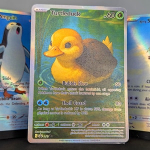Pode incluir: Um cartão Pokémon holográfico Turtleduck com uma criatura amarela parecida com um pato na água. O cartão tem o texto "Turtleduck" e "Bubble Blow" com detalhes de ataque. Outros cartões Pokémon são visíveis no fundo.