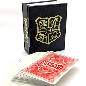 May include: A black playing card box with gold lettering and a crest, next to a deck of playing cards. The box is shaped like a book and has the words "Harry Potter" printed on the side. The playing cards have a red design and the words "Harry Potter" printed on them.