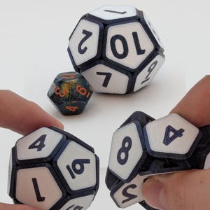 May include: Three polyhedral dice are displayed against a white background. Two large dice are white with black numbers and black borders. A smaller, multi-colored die is also present. The dice are used for tabletop games.