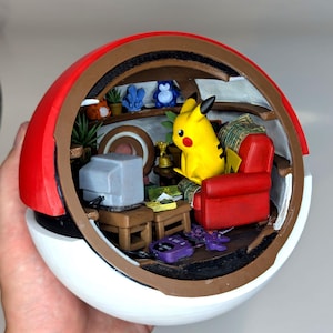 May include: A detailed diorama inside a Poké Ball featuring a yellow Pikachu figure in a red armchair. The scene includes a vintage TV, small tables, and miniature plants, all within a red, brown, and white sphere.