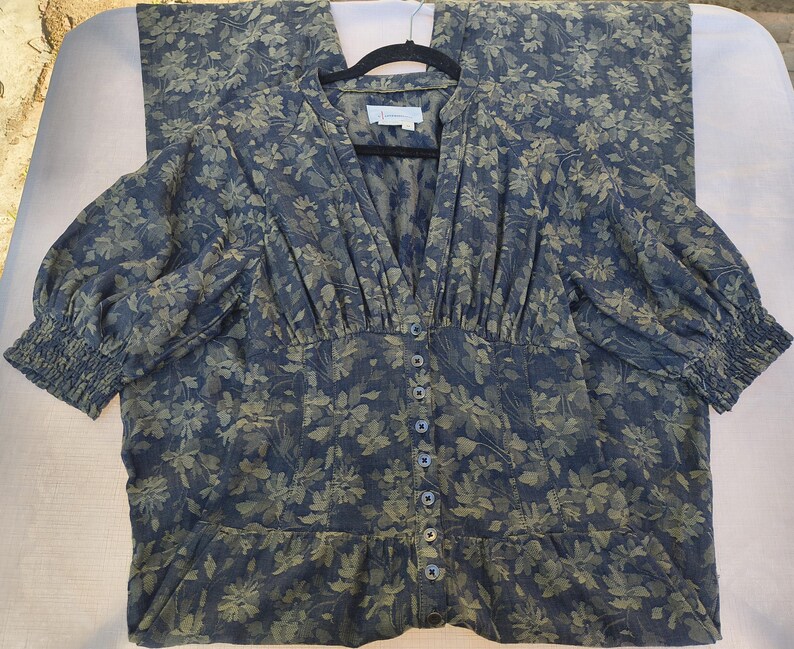 May include: A blue floral print dress with a V-neckline and button-down front. The dress features three-quarter sleeves with elastic cuffs and a fitted waist. The fabric has a pattern of green and blue floral designs.