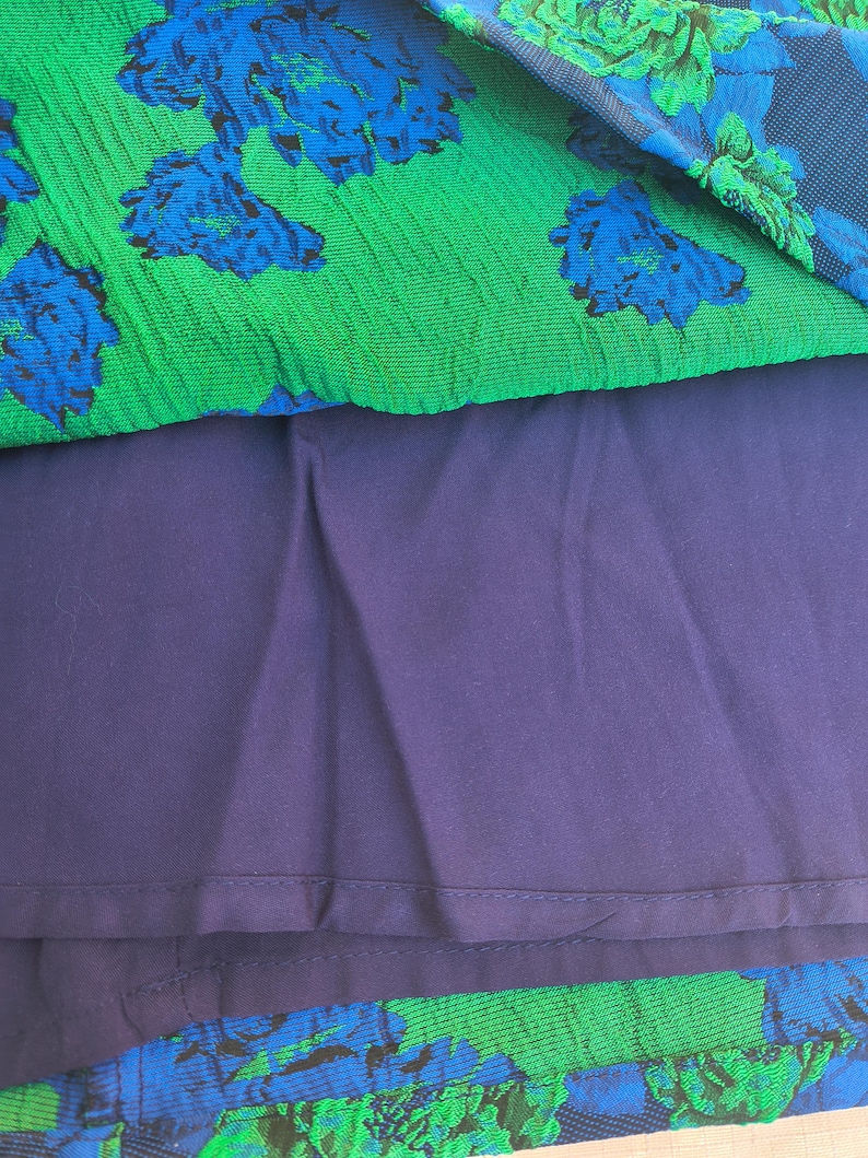 May include: A close-up of a dress with a layered design. The top layer is green with a blue floral pattern. The middle layer is a solid dark purple. The bottom layer is a dark purple floral pattern.