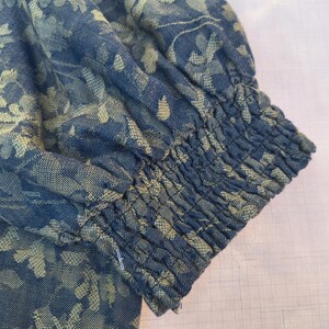 May include: Close-up of a blue fabric sleeve with a floral pattern in a lighter shade of blue. The sleeve has gathered elastic at the wrist. The fabric appears to be a woven material.