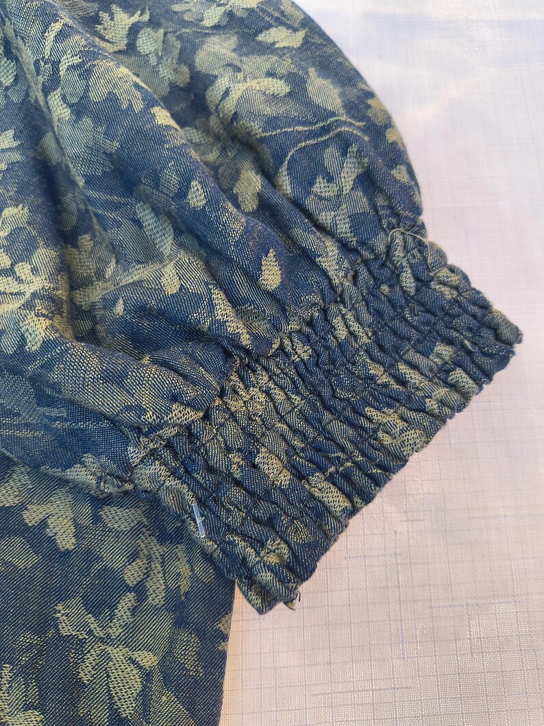 May include: Close-up of a blue fabric sleeve with a floral pattern in a lighter shade of blue. The sleeve has gathered elastic at the wrist. The fabric appears to be a woven material.