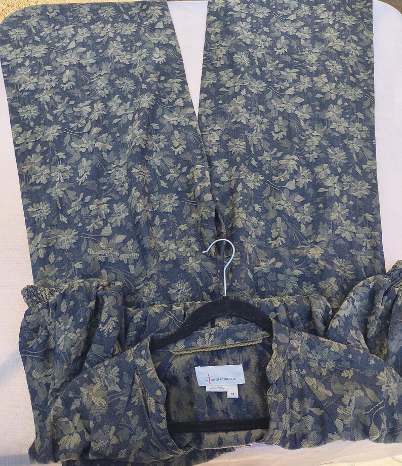 May include: A two-piece floral print outfit in shades of blue and green. The set includes wide-leg pants and a button-down shirt. The shirt is hanging on a black hanger. The fabric has a delicate floral pattern.
