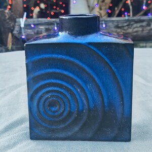 May include: A square, ceramic vase with a dark blue exterior and a spiral design in lighter blue. The vase has a rectangular opening at the top and sits on a light-colored surface. The background is blurred with colorful lights.