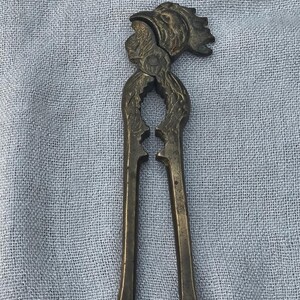 May include: Antique brass nutcracker with a detailed rooster head design. The nutcracker features long handles and a hinged mechanism for cracking nuts. The brass has a weathered appearance, suggesting age and use. A decorative kitchen tool.