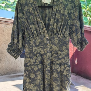 May include: A dark green floral print dress with a V-neckline and button-down front. The dress features three-quarter sleeves with elastic cuffs and a fitted waist. The fabric has a subtle sheen, and the floral pattern is in shades of green and navy.