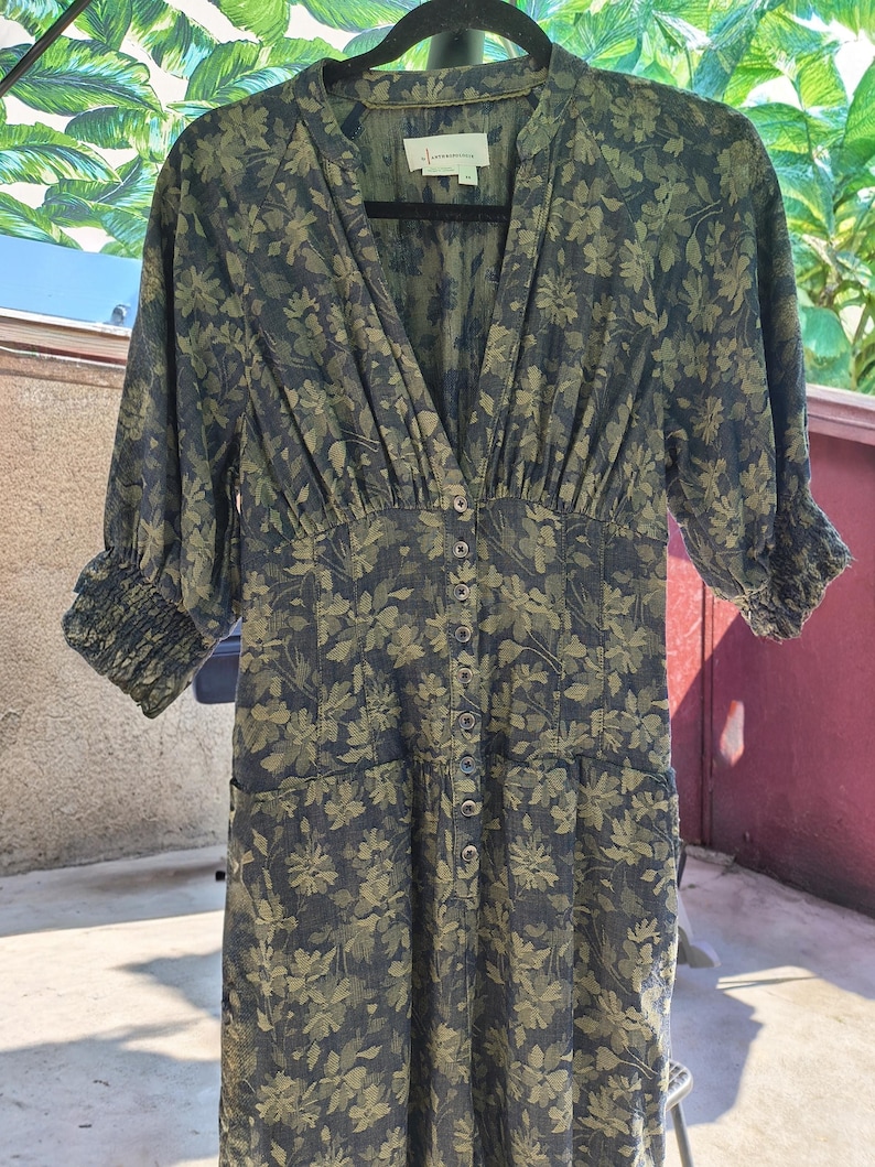 May include: A dark green floral print dress with a V-neckline and button-down front. The dress features three-quarter sleeves with elastic cuffs and a fitted waist. The fabric has a subtle sheen, and the floral pattern is in shades of green and navy.
