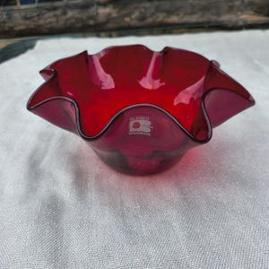 May include: A ruby red glass bowl with a ruffled edge. The bowl has a circular shape with a wavy, petal-like rim. The base of the bowl is smooth, and the glass appears to be transparent. The bowl has a sticker that says "Blenco Handmade".