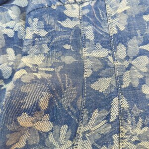 May include: Close-up of blue denim fabric with a white floral pattern. The fabric is stitched with white thread, and the floral design is a repeating pattern of leaves and flowers. The overall look is casual and summery.