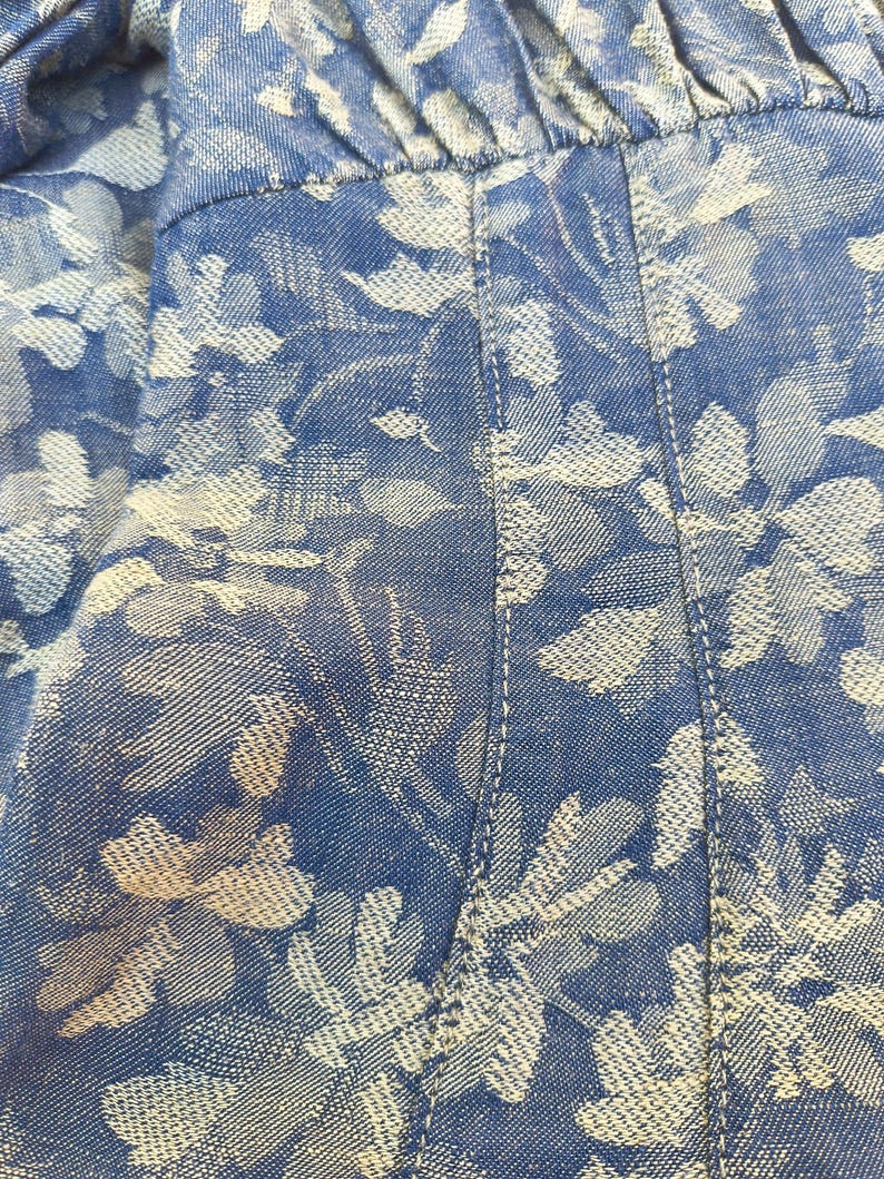 May include: Close-up of blue denim fabric with a white floral pattern. The fabric is stitched with white thread, and the floral design is a repeating pattern of leaves and flowers. The overall look is casual and summery.