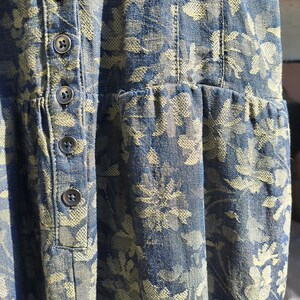 May include: Close-up of a blue denim dress with a floral pattern in a lighter shade. The dress features a button-down front and a gathered waist. The fabric has a textured appearance.