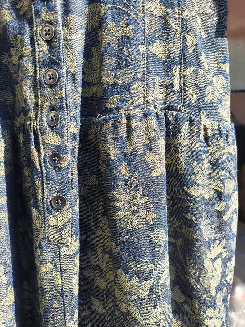May include: Close-up of a blue denim dress with a floral pattern in a lighter shade. The dress features a button-down front and a gathered waist. The fabric has a textured appearance.