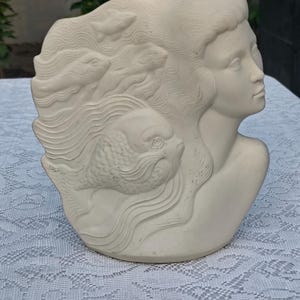 May include: A matte off-white ceramic bust sculpture featuring the profile of a face with long flowing hair that transitions into stylized waves and three distinct fish figures integrated into the design, creating a cohesive relief texture.