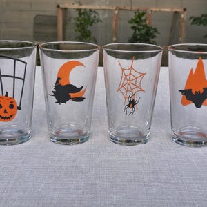 May include: Four clear glass tumblers with Halloween-themed designs. Each glass features a different orange and black graphic: a jack-o'-lantern, a witch on a broom, a spider and web, and a bat with mountains.