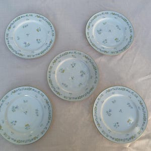May include: Five white ceramic plates arranged in a circular pattern. Each plate features a delicate rim and center decoration consisting of small, scattered blue and yellow floral sprigs. The plates have a smooth, glossy finish and a circular shape.