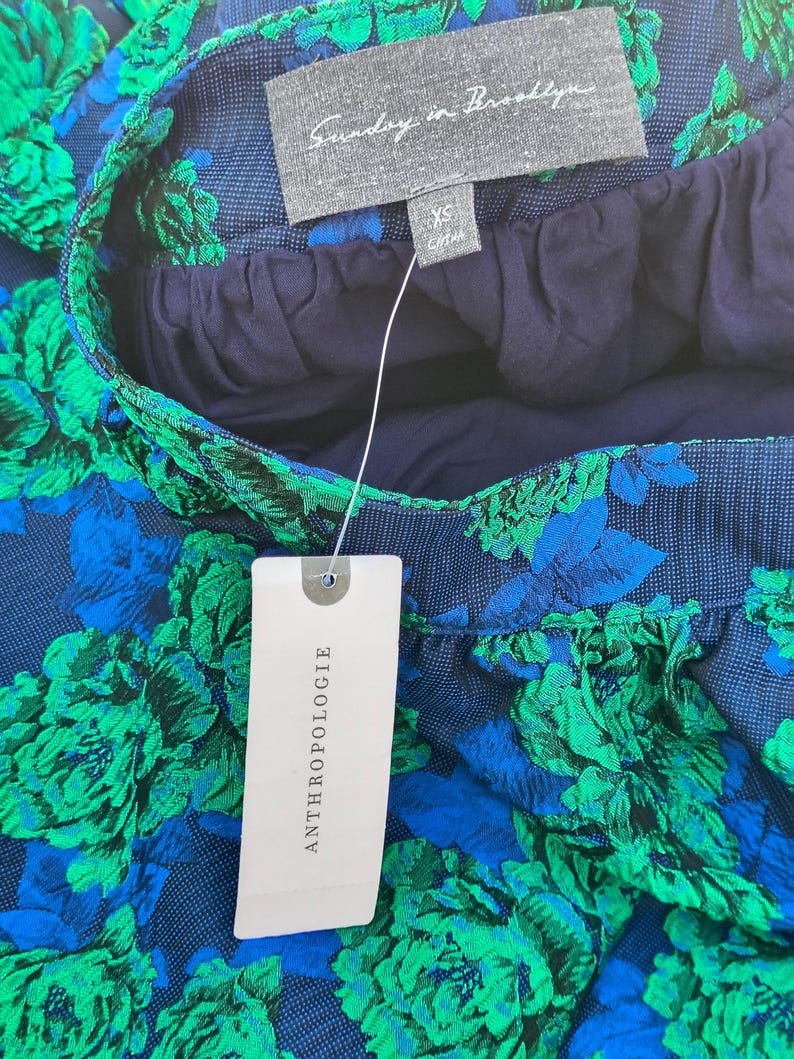 May include: Close-up of a skirt with a floral pattern in shades of blue and green. The skirt has a navy blue waistband and lining. A tag from Anthropologie is attached. The label reads "Sunday in Brooklyn".