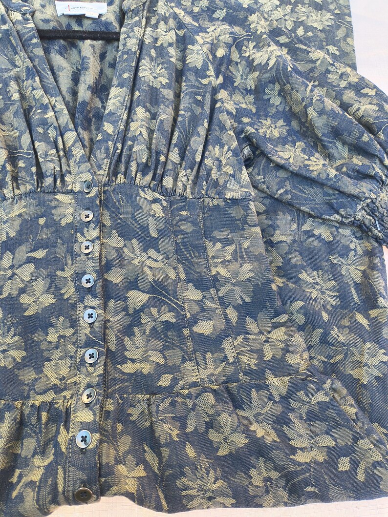May include: A blue floral print dress with a V-neckline and button-down front. The dress features a fitted bodice, long sleeves, and a ruffled hem. The floral pattern is in shades of beige and blue.