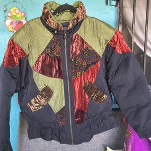 May include: A vintage, multi-colored jacket with a high collar and a front zipper. The jacket features panels of olive green, burgundy, and black fabric, with decorative paisley and velvet accents. The jacket has a cinched waist and long sleeves.