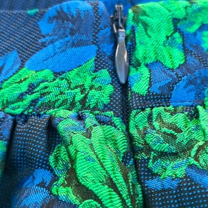 May include: Close-up of a fabric with a floral pattern in shades of blue and green. The design features a textured weave with a silver zipper. The fabric appears to be part of a garment, possibly a skirt or dress.