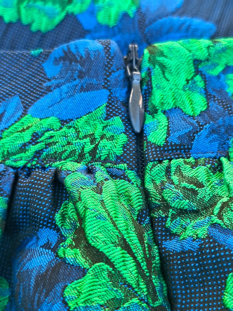 May include: Close-up of a fabric with a floral pattern in shades of blue and green. The design features a textured weave with a silver zipper. The fabric appears to be part of a garment, possibly a skirt or dress.