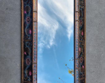 May include: A rectangular mirror with a decorative frame. The frame is made of wood and has a dark blue, pink, and gold paint design. The mirror reflects a blue sky with white clouds.