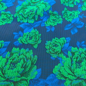 May include: Close-up of a fabric with a floral pattern. Large green flowers with detailed textures are set against a dark blue background. Blue leaves and stems are interspersed among the flowers, creating a vibrant contrast.