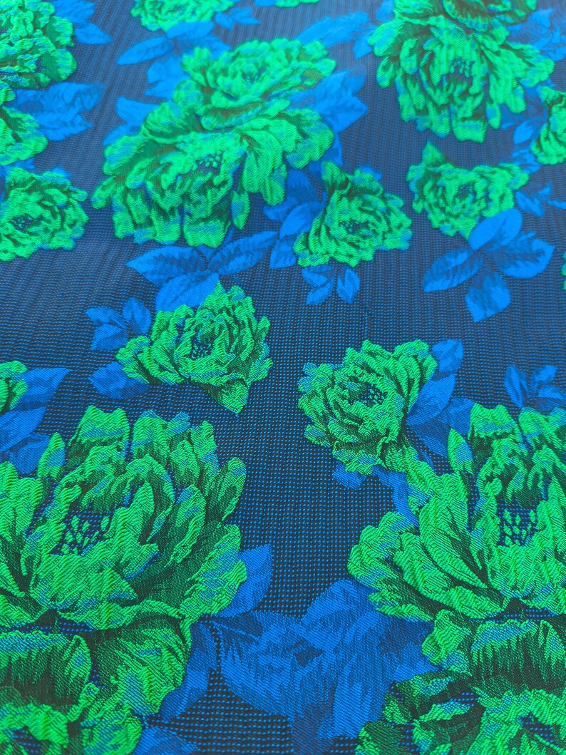 May include: Close-up of a fabric with a floral pattern. Large green flowers with detailed textures are set against a dark blue background. Blue leaves and stems are interspersed among the flowers, creating a vibrant contrast.