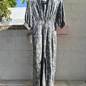 May include: A blue floral print jumpsuit with a V-neckline and button-down front. The jumpsuit has short sleeves, a fitted waist, and wide legs. The fabric has a light blue background with a yellow floral pattern. The jumpsuit is hanging on a black hanger.