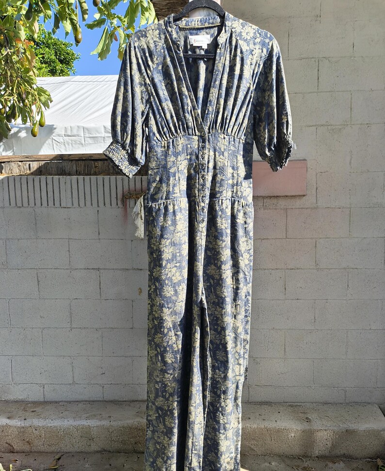 May include: A blue floral print jumpsuit with a V-neckline and button-down front. The jumpsuit has short sleeves, a fitted waist, and wide legs. The fabric has a light blue background with a yellow floral pattern. The jumpsuit is hanging on a black hanger.