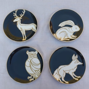 May include: Set of four decorative ceramic plates with animal designs. Each plate features a dark blue background with a gold rim and a white and gold illustration of a deer, rabbit, owl, and fox. These plates are perfect for serving appetizers or as decorative pieces.