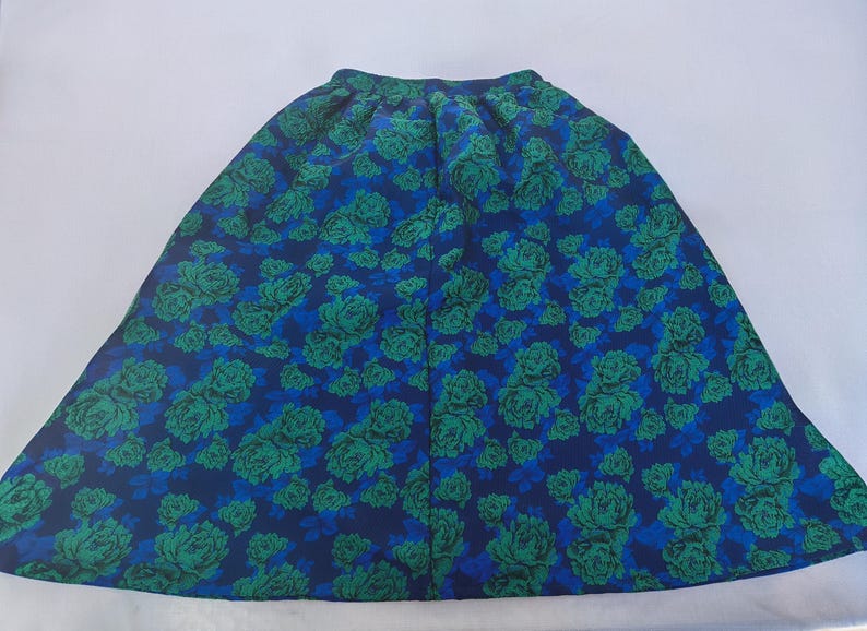 May include: A full-length skirt with a vibrant floral pattern. The skirt features a deep blue background adorned with large green and teal rose blooms. The fabric is gathered at the waist, creating a flowing silhouette.