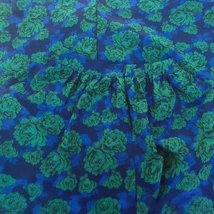 May include: A close-up of a garment with a floral pattern. The fabric features a repeating design of green roses on a dark blue background with lighter blue accents. The garment has a gathered waistline.