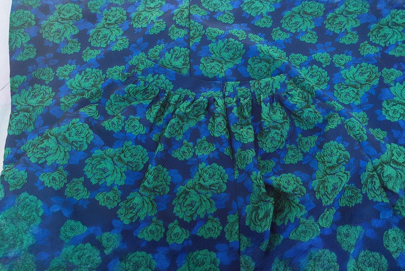 May include: A close-up of a garment with a floral pattern. The fabric features a repeating design of green roses on a dark blue background with lighter blue accents. The garment has a gathered waistline.
