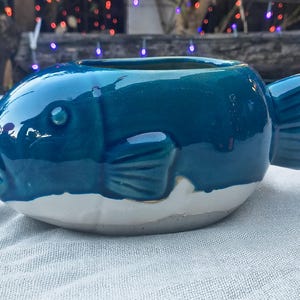 May include: A ceramic planter shaped like a blue fish with a white base. The fish has detailed fins and a wide open mouth. The planter is glazed with a glossy finish, and is suitable for small plants or succulents.