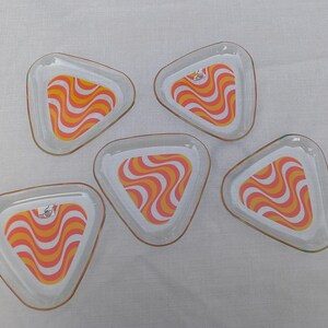 May include: Five clear, triangular glass dishes with a retro orange and white wavy pattern. Each dish has a thin, gold-colored rim. The dishes are arranged on a white surface.