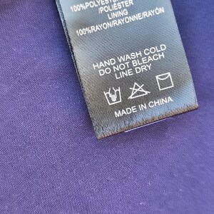 May include: Close-up of a black clothing tag with white text. The tag reads "SELF 100% POLYESTER/POLYESTER /POLI&Eacute;STER LINING 100% RAYON/RAYONNE/RAY&Oacute;N HAND WASH COLD DO NOT BLEACH LINE DRY MADE IN CHINA". The tag is on a purple fabric.