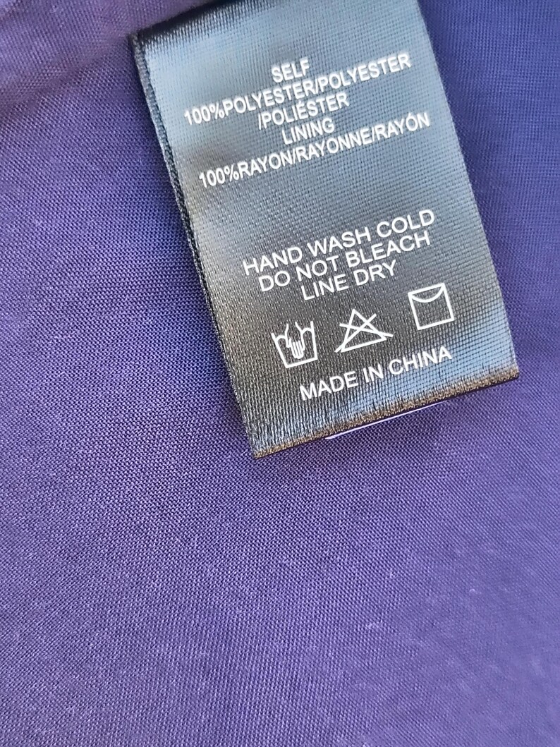 May include: Close-up of a black clothing tag with white text. The tag reads "SELF 100% POLYESTER/POLYESTER /POLI&Eacute;STER LINING 100% RAYON/RAYONNE/RAY&Oacute;N HAND WASH COLD DO NOT BLEACH LINE DRY MADE IN CHINA". The tag is on a purple fabric.