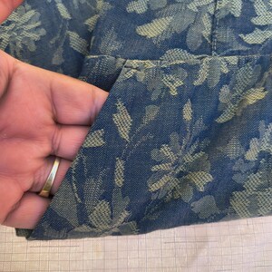 May include: Close-up of a blue denim fabric with a floral pattern in a lighter shade of blue. The fabric is folded, revealing a pocket. The texture of the fabric is visible, and the overall look is casual and stylish.
