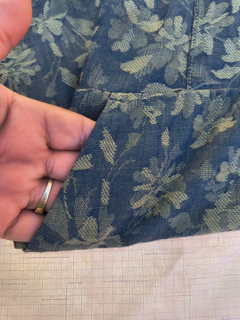 May include: Close-up of a blue denim fabric with a floral pattern in a lighter shade of blue. The fabric is folded, revealing a pocket. The texture of the fabric is visible, and the overall look is casual and stylish.