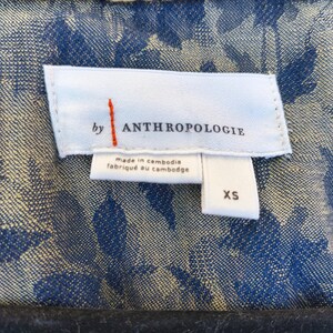 May include: Close-up of a blue floral patterned jacket with a white label that reads "by ANTHROPOLOGIE" with the text "made in cambodia" and "fabriqu&eacute; au cambodge". The size "XS" is also visible.