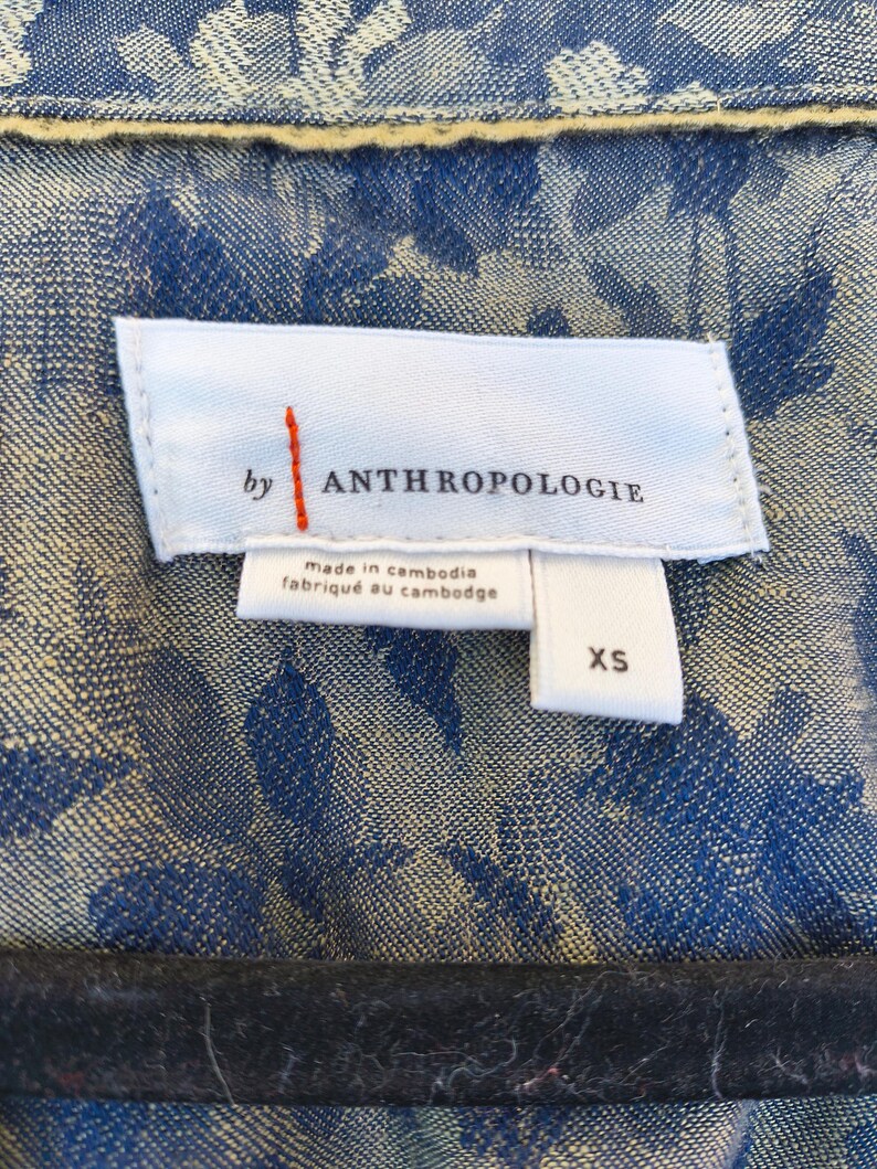 May include: Close-up of a blue floral patterned jacket with a white label that reads "by ANTHROPOLOGIE" with the text "made in cambodia" and "fabriqu&eacute; au cambodge". The size "XS" is also visible.