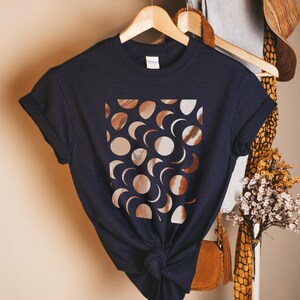 Könnte beinhalten: Ein marineblaues T-Shirt mit einem quadratischen Design aus Mondphasen in Braun- und Weißtönen. Das T-Shirt ist am unteren Ende geknotet und hängt an einem Holzbügel. Teil einer Bekleidungskollektion.