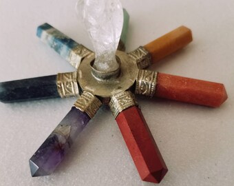 May include: A star-shaped crystal grid featuring seven distinct gemstone points radiating from a central metal hub. The points include purple amethyst, dark black stone, blue stone, orange, red, and dark red stones, with a clear quartz point in the centre.
